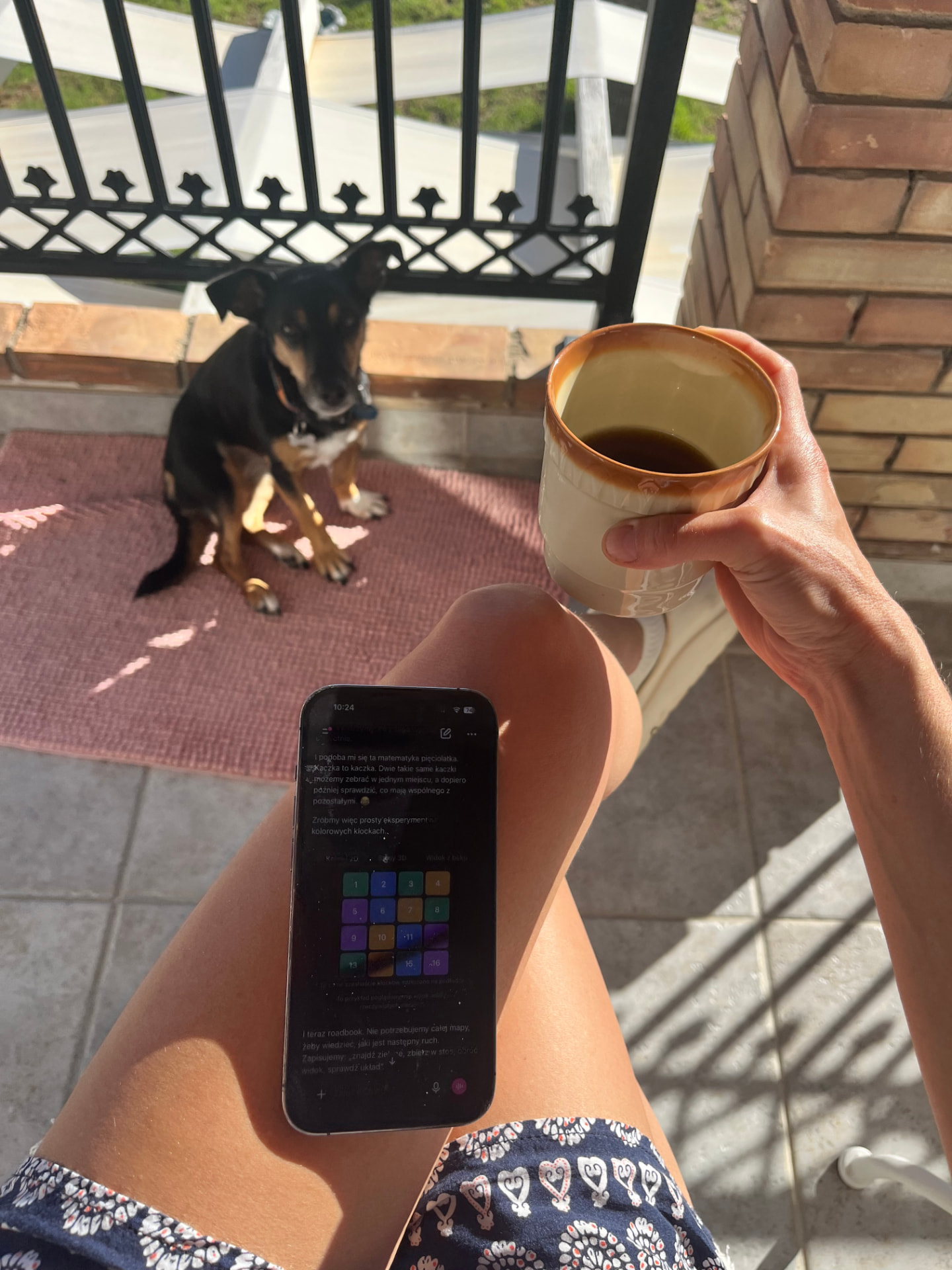 A sunny morning seen from Anna's own point of view: a cup of coffee in her hand, her phone on her lap, and Bułka sitting on the rug in front of her